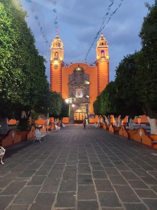 Orange colonial church with twin bell towers in San Pedro Cholula at dusk, flanked by green trees